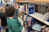 Two women stand in a library aisle in front of shelves of books. One visible title reads "Own Your Period."