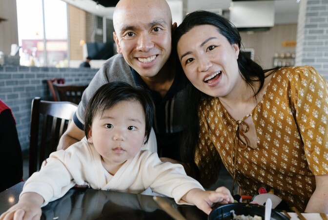 An Asian American family of three poses for the camera at a restaurant. From left to right: a year-old baby, a 40-something Filipino American man and a Korean American woman.