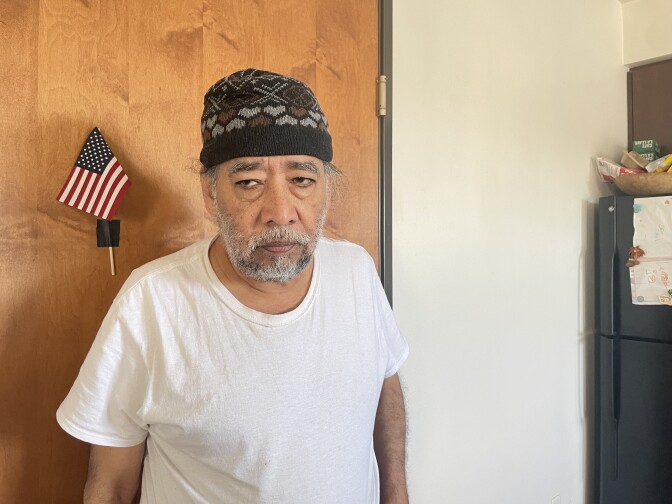 A man with gray hair, mustache, and beard poses for a photo. He looks away from the camera. There is a refrigerator with children's art and a U.S. flag taped to a door in the background. 