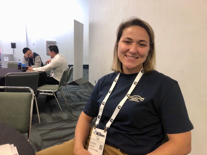 A woman with light skin tone and short blonde hair wears a blue shirt and smiles at the camera while sitting at a table in a conference room. 