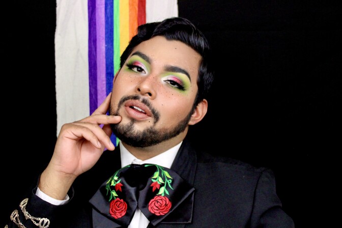 A man with a goatee wears a black suit with a black bowtie embroidered with red flowers. He has green, pink and silver eyeshadow.