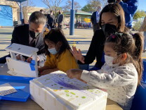 Two adults interact with two young children who are decorating and building with styrofoam blocks at an outdoor work table in a school play yard. All in the picture are wearing face coverings to prevent COVID-19 spread.