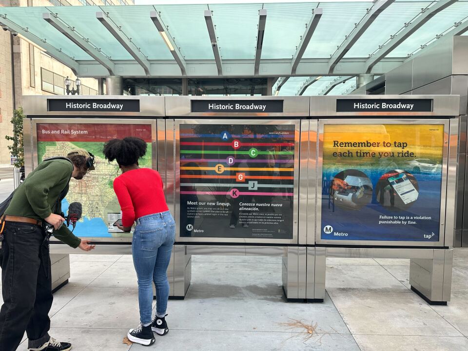 A fair-skinned man in black pants and a forest green top with headphones and a mic examines a bus map with a young Black woman in jeans and a red shirt. 