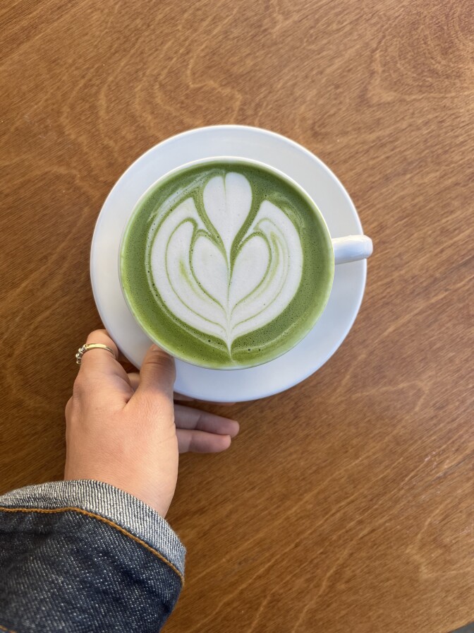 a white ceramic cup and saucer sit on a brown wooden surface. The cup is filled with a green liquid with white latte art in the center of a heart design. 