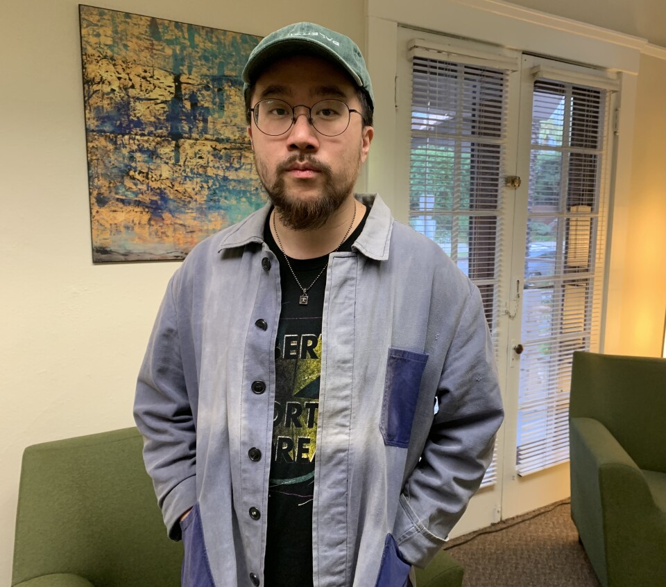 A Chinese American man in his 30s wears a denim shirt, eyeglasses and a solemn look on his face.