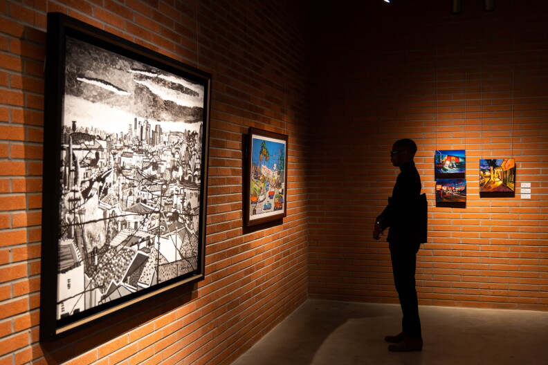 A man in shadows stands looking at art hanging on the walls, including a large black and white piece in front, a more colorful piece next to it, with several more colorful pieces on the brick wall to the man's side.