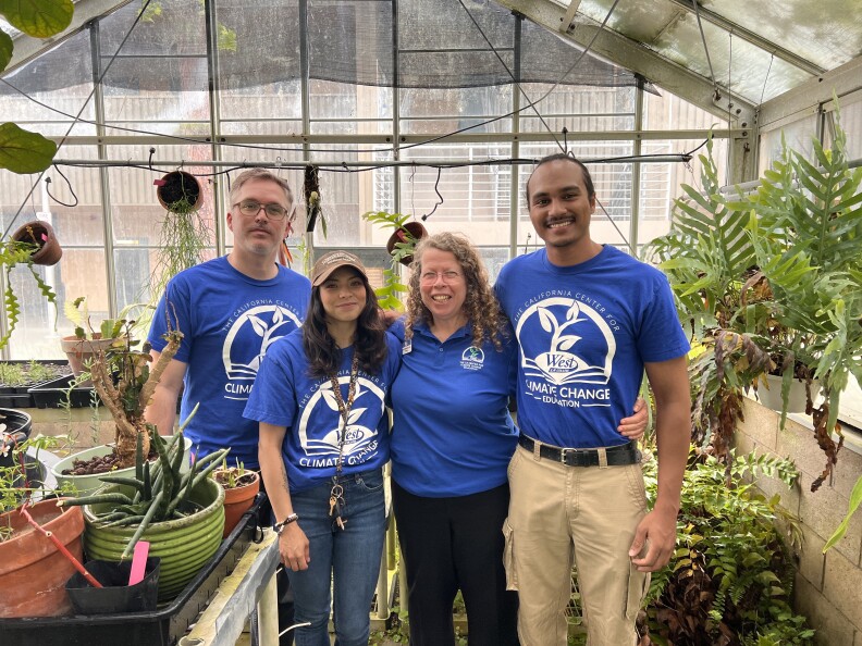Four adults with various skin tones and wearing blue shirts stand in a greenhouse surrounded by plants.