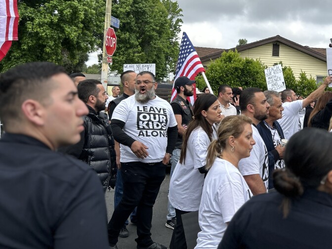 A man with a medium-light skin tone stands in a crowd of protesters on the street wearing a black and white shirt that says leave our kids alone. In the foreground are police officers.