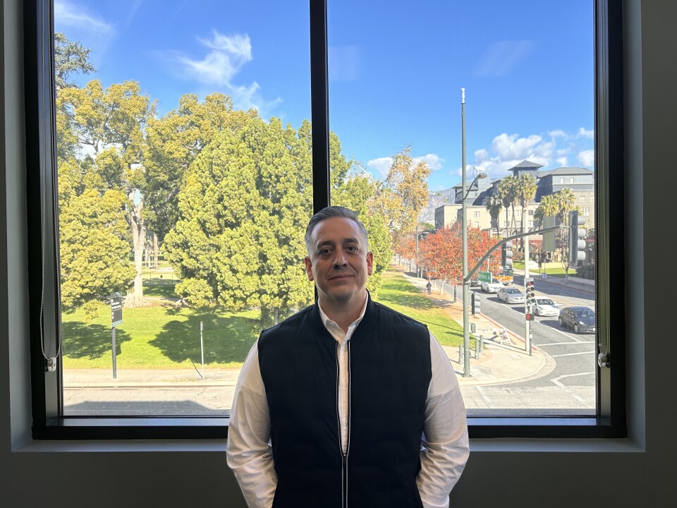 A man wearing a white long-sleeve button down under a black vest is standing in front of a window overlooking a green park and mountains in the distance. 