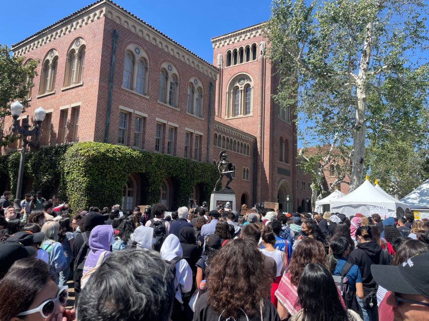 A large group of people with their backs mostly towards the camera stand in front of USC's Tommy Trojan statue. A red brick building with beige windows is visible in the background.
