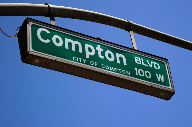 A close-up of a green street sign hanging from a lamp post with a blue sky in the background. The sign reads "Compton Blvd 100 W City of Compton"