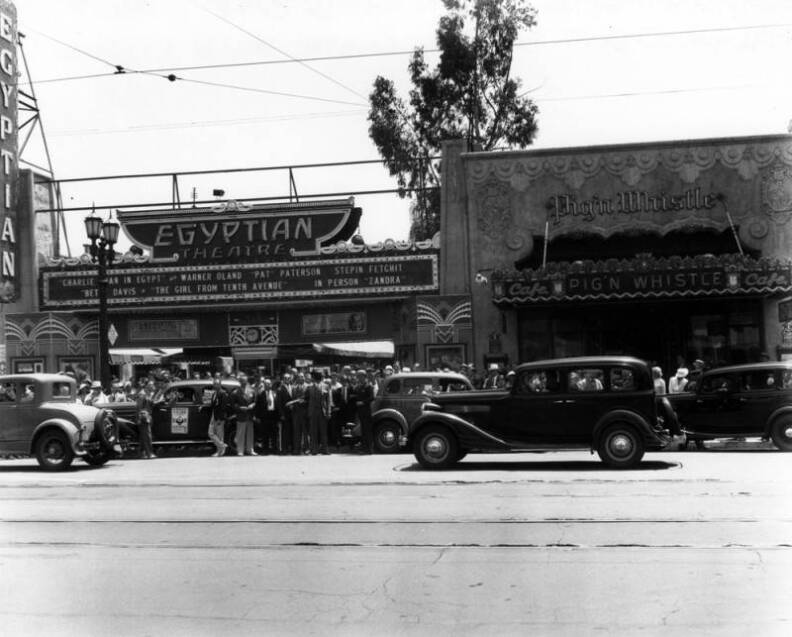 A crowd has gathered in the street and on the sidewalk around a car with a sign, "Magical No-Jinks."