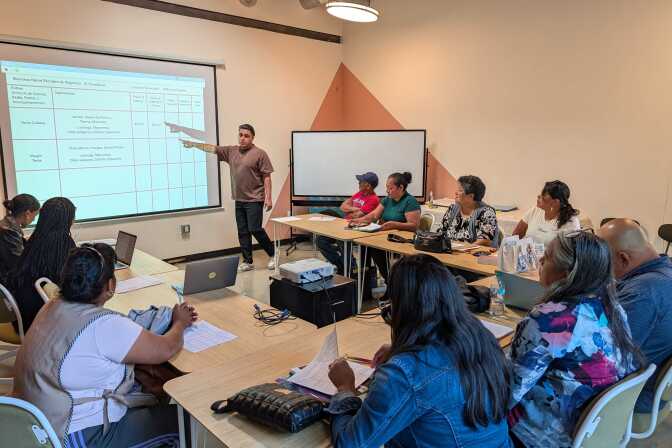 Ten people sit in a classroom. They look at a person standing, pointing to an image on a screen.