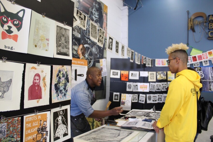 two men stand at a table stacked with prints and talk. prints hang all around them.
