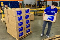 A male presenting person carries a cardboard box. A stack of about 30 boxes is next to him.