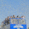 A double-decker bus and the people celebrating on top are nearly obscured by a cloud of confetti. The front of the bus reads "World Champions."
