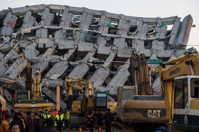 A general view shows excavator vehicles and rescue workers in front of a building (background) which collapsed in the 6.4 magnitude earthquake, in the southern Taiwanese city of Tainan early on February 9, 2016. Rescuers are set to start using diggers and extractors to remove giant concrete slabs once they have ensured all residents from the upper parts of the rubble have been freed.  AFP PHOTO / ANTHONY WALLACE / AFP / ANTHONY WALLACE        (Photo credit should read ANTHONY WALLACE/AFP/Getty Images)