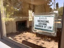 A front window in a home, with the bare living room and fireplace seen inside. A black and white sign that reads "Warning state property trespassing, loitering forbidden by law" is taped on the front facing out towards the street.