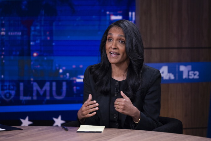 A woman with black hair past her shoulders sits at a news desk gesturing while speaking, with LMU and station logos on screens behind them.