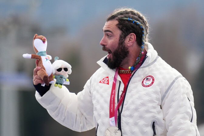 A man with long hair in a braid, wearing a white puffer jacket, smiles as he holds two stuffed animals