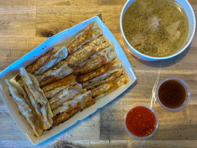Pan-fried dumplings fill a takeout box next to a bowl of broth soup and two dipping sauces, one red and one brown.