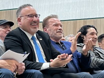A man with glasses and a blue tie and another man wearing a blue suit sit in a theater and applaud.