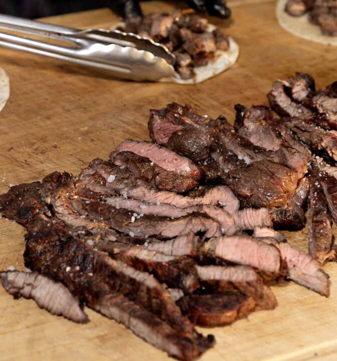 Slices of grilled steak with charred edges and pink centers, sprinkled with coarse salt, on a wooden cutting board, with tongs and a taco in the background.
