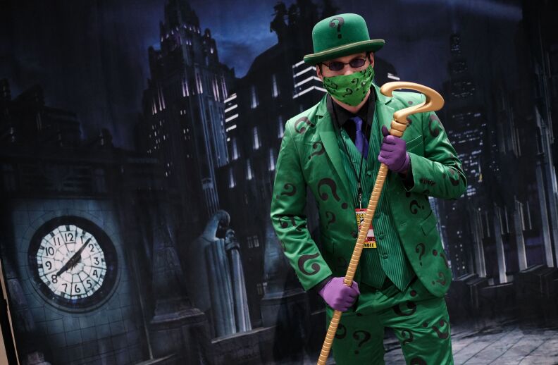 A cosplayer wearing a green riddler costume, including a green face mask with question marks on it, stands in front of a Gotham City backdrop featuring skyscrapers, ominous statues, and a clocktower. He holds a staff with a question-mark-shaped top.