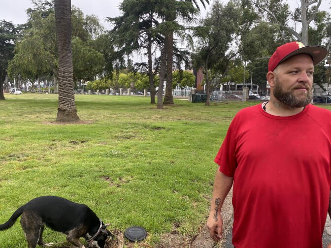 A tall bearded white man in a red baseball cap and red T-shirt stands on a lawn in front a grove of trees at the West Los Angeles VA campus. A small dog sniffs the grass behind him.