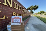 Four medium-sized cardboard boxes sit on a red brick structure. On top of the boxes is a laminated sheet of paper that reads, "Hilda L. Solis Operation Laptops." Off to the right is a sidewalk. 