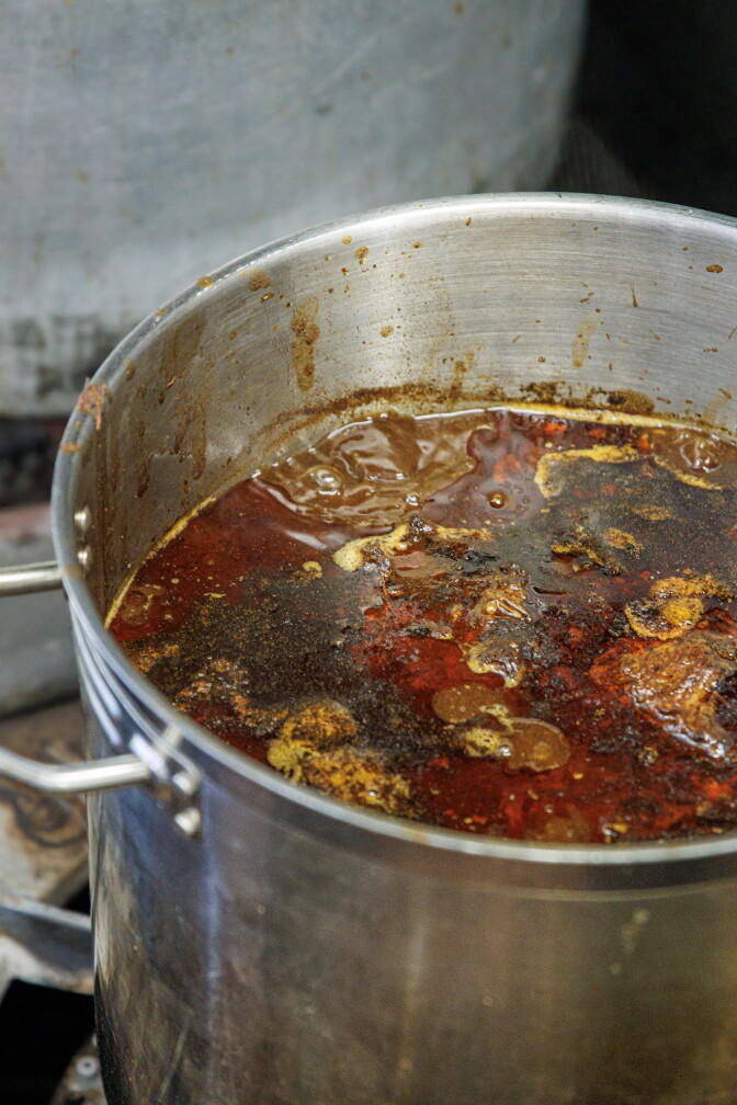 A large stainless steel stockpot filled with deep red, chile-spiced braising liquid, with a layer of rendered fat and pieces of beef visible at the surface.
