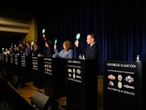 A group of candidates in suits stand on a stage in a long line side by side, each in front of a podium. One podium, which has the name "George Gascón" printed on the front, is empty.