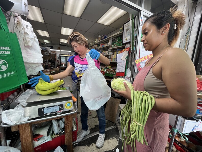 An Asian woman in a pink, yellow and white shirt bags bananas while a Latina customer waits holding cabbage and yardlong beans.