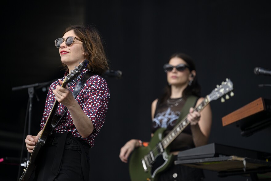 Carrie Brownstein stands on stage, performing during a Sleater-Kinney set. She's wearing a shirt with blue, white, and read hearts and playing a guitar. Another band member is holding a green guitar on stage out of focus behind her.