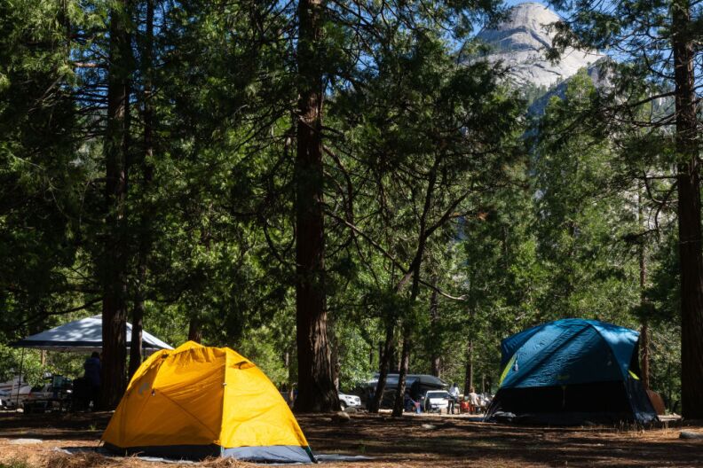 An orange tent, a white pop up and a blue tent are seen at a campground. Trees surround the tents and a mountain can be seen in the background
