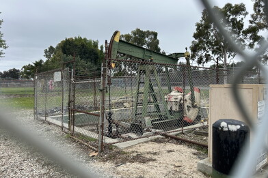 A green pumpjack surrounded by a chainlink fence under cloudy skies. 