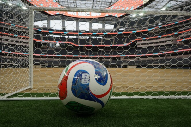 A multi-colored soccer ball sits on a patch of grass and beside a net. In the background is an empty stadium.