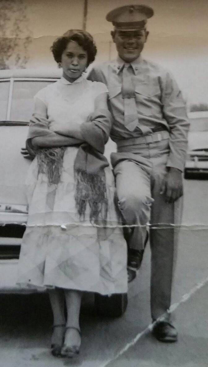 A male presenting person dressed in a military uniform and a female presenting person in a dress in front of a car.