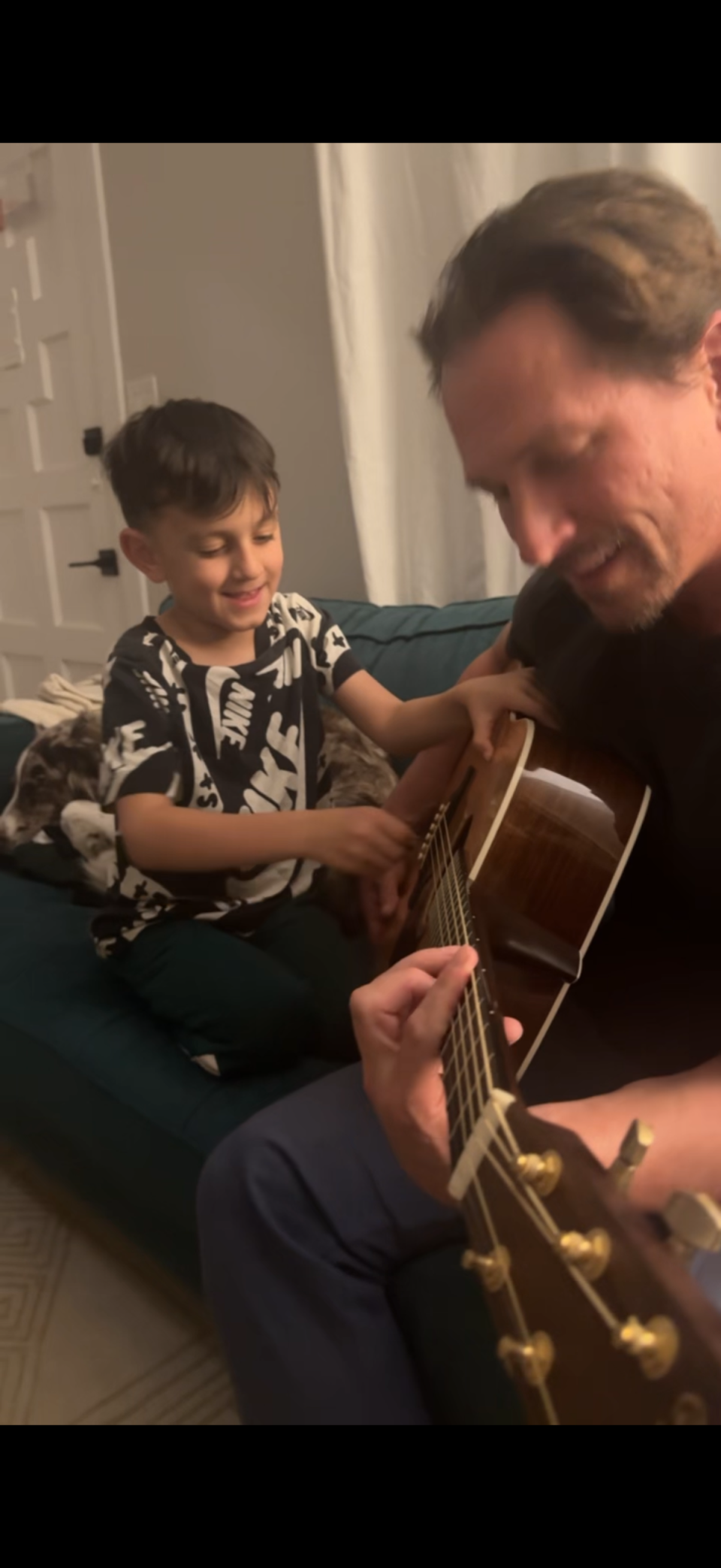 A man forms a chord on the fret board of an acoustic guitar, while a boy touches the strings.