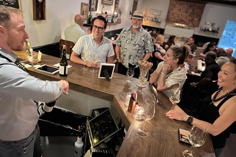 A group of people gather around a bar while the waiter pops open a bottle of wine.