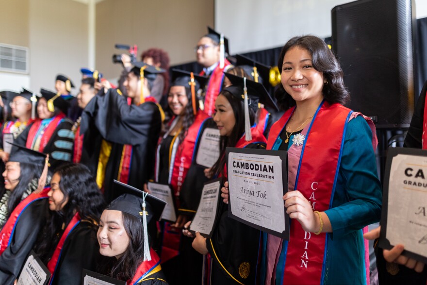 A young Cambodian woman with wavy black hair stands among a crowd of graduates posing on indoor bleachers. She is holding a certificate that reads "Cambodian Graduation Celebration" and her name, "Ariya Tok." She has no cap and gown but wears a blue dress or tunic with a pink sash decorated with a flower. A formal red graduation sash is draped over that, hanging from from both shoulders.