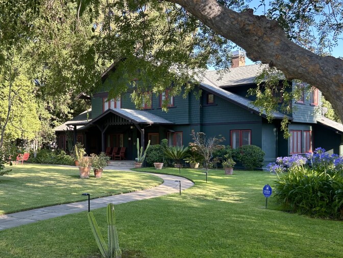 A dark green house with red window paneling is surrounded by bushes. There is a green front lawn and a cement path leads to the home's front door. A large tree sits out front. 