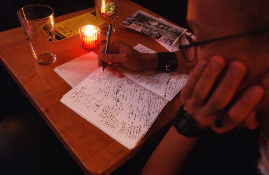 A close-up of a young adult in profile, who appears to be Brown or Black, sitting at a dimly lit table at a bar writing in a notebook. The table also has an empty beer glass and a small candle, and a napkin, menu and flyer.
