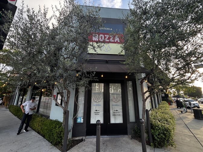  The corner building, which features a tri-colored signage that reads "Osteria Mozza," is painted light blue and has black awnings. Its entrance includes windowed double doors. To the left of the building stands a man on the sidewalk, wearing a white short-sleeved shirt, a baseball cap, and sunglasses.