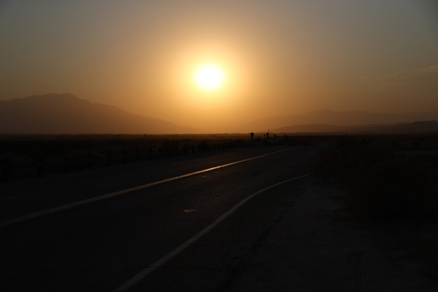 A deep orange sun sets in a hazy sky with a desert scape and mountains in the foreground.