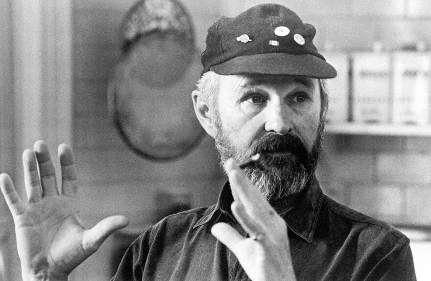 A bearded white man in a cap motions with his hands while looking off camera.