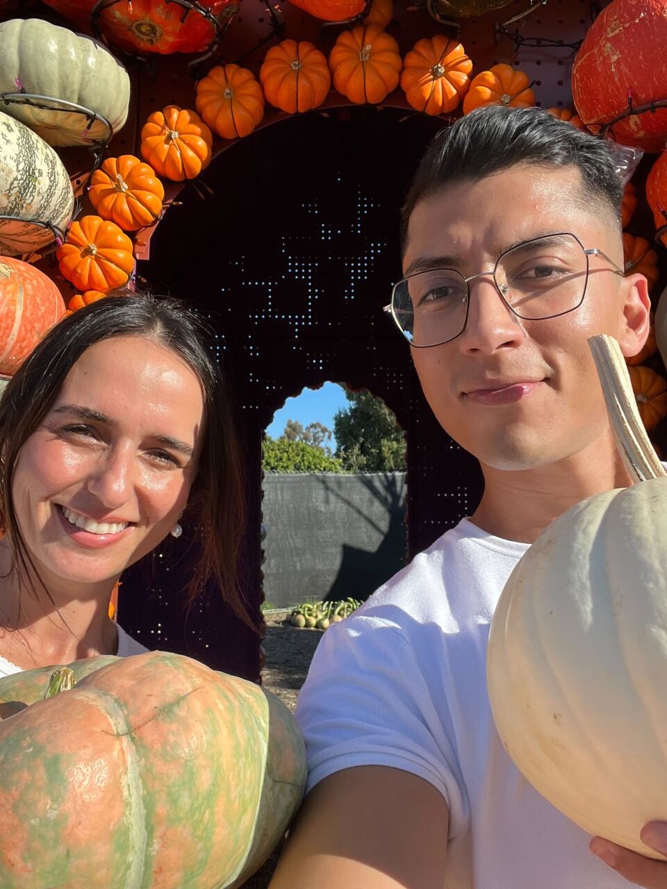 A white woman with brown hair and a Latino man with glasses smile at the camera holding up pumpkins 