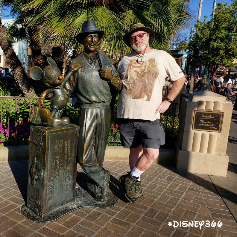 A man with light-tone skin and gray beard wears a T-shirt, sun hand and sunglasses as he leans on a bronze stautude of Walt Disney with a small Mickey Mouse.