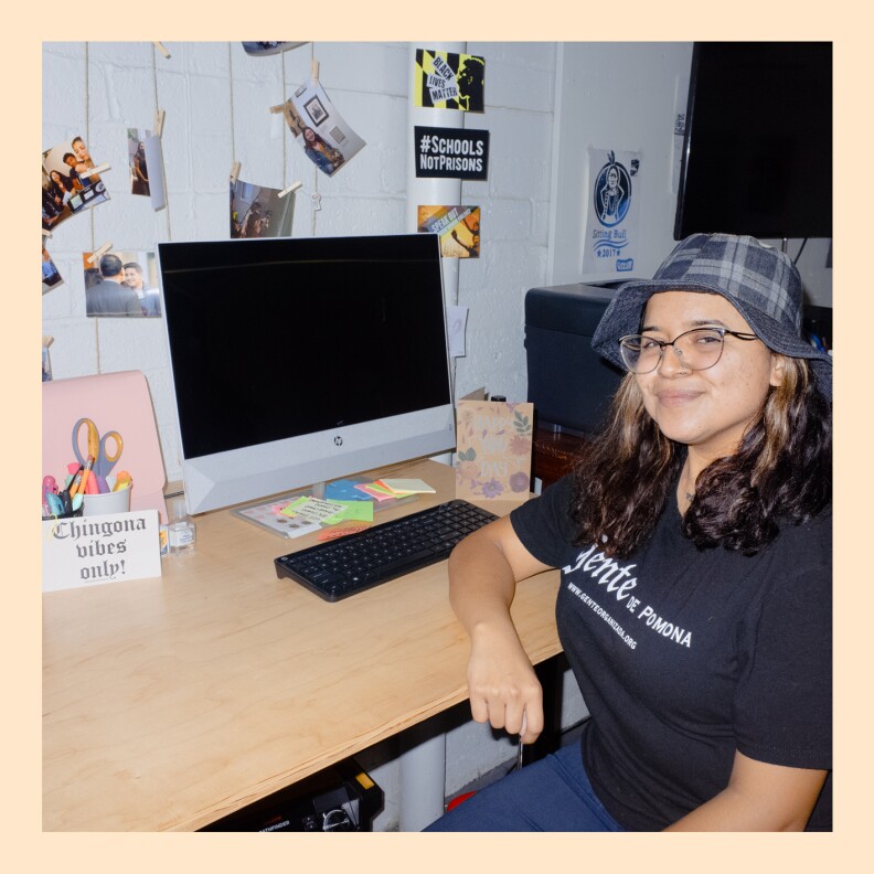 A young girls sits in front of a computer at a desk. She wears a shirt that reads "gente de pomonoa." On the wall around her computer various photos and signs are on display. On the desk, there is a sign that reads "Chingona vibes only!"