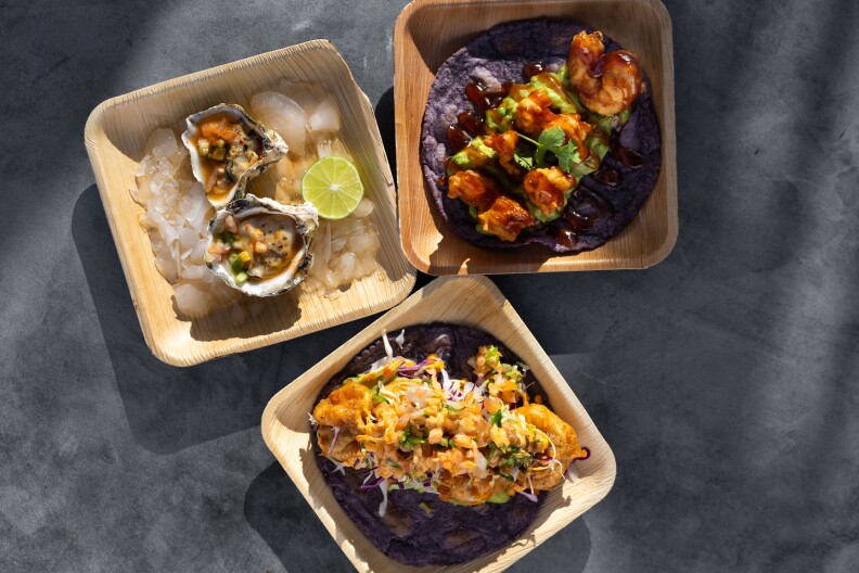 Overhead photo of three bamboo plates: One plate holds two oysters on the half shell, sitting on ice, alongside a slice of lime. A second plate holds a blue corn tortilla brimming with shrimp and avocado. The third plate holds a blue corn tortilla topped off with battered shrimps and traditional toppings.